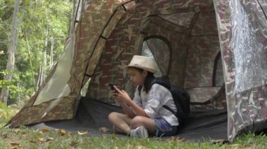 Asian cute girl with backpack sitting and using mobile phone in front of the tent in natural park area during camping.