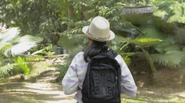 Rear view of asian girl with backpack walking in tropical natural park in thailand.