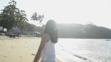 Asyalı şirin kız hasır şapka takıyor ve günışığı altında kollarını kumsalda sallıyor. Yavaş çekim.
