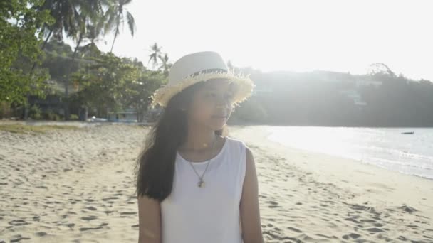 アジアのかわいい女の子は 朝の日差しの下でビーチを歩くわらの帽子とカジュアルなドレスを着ています スローモーション — ストック動画