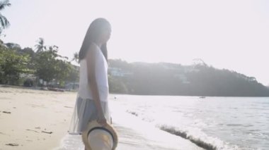 Asyalı şirin bir kız, sabahleyin plajda güneş ışığı altında günlük kıyafet giymiş hasır şapka takıyor. Yavaş çekim.