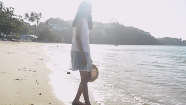 アジアのかわいい女の子は 午前中に日光の下でビーチに立ってカジュアルなドレスを着てわら帽子を保持します スローモーション — ストック動画