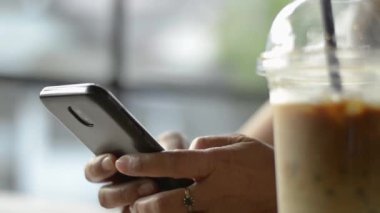 Asyalı bir kadın, kahve dükkanında bir bardak soğuk içecek içerken iletişim kurmak ve sosyal medyada sohbet etmek için cep telefonundan akıllı mesajlar atıyor..