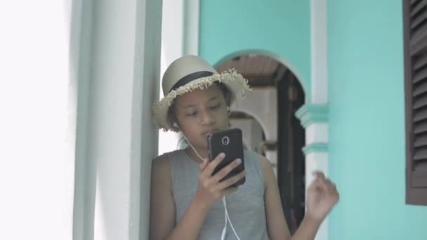 在泰国普吉岛的奇诺葡萄牙风格建筑中 身穿休闲装 头戴草帽 头戴手机和耳机的亚洲可爱女孩站在拱廊边 禁止慢动作射击 — 图库视频影像
