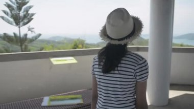 Sıradan giyinmiş, hasır şapkalı Asyalı tatlı bir kız. Manzara kulesinde yürüyor ve Pofet sahilinin yukarısındaki büyüleyici manzaranın keyfini çıkarıyor. Güzel kız şapkasını çıkarır ve kollarını havaya kaldırır. Yavaş çekim.