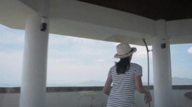 Sıradan giysili ve hasır şapkalı Asyalı tatlı kız Manzara kulesine doğru koşuyor ve Pofet sahilinin yukarısındaki büyüleyici manzaranın keyfini çıkarıyor. Güzel kız şapkasını çıkarır ve kollarını havaya kaldırır. Yavaş çekim.