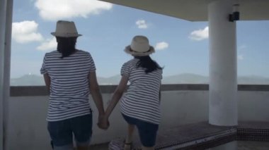 Anne ve kızı saman şapka takıyor, sıradan bir elbise giyiyorlar. Manzara kulesine doğru yürüyorlar ve Pofet Körfezi 'nin üzerindeki büyüleyici manzaranın keyfini çıkarıyorlar. Anne ve kızı mutlu bir şekilde kollarını havaya kaldırıyor. Arka görüş takip çekimi. Yavaş çekim.