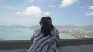Küçük kızın, yaz boyunca Phuket 'te, körfezin manzaralı güzel manzarasını izlerken annesine mutlu bir şekilde sarılışının dikiz görüntüsü. Aşkın ifadesi.