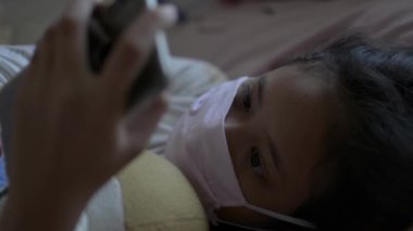 Asyalı sevimli kız koruyucu maske takıyor ve yatakta yatarken cep telefonu kullanıyor. Corona virüs koruması. Covid 19 salgını. Sağlıklı ve güvenli yaşam konsepti.