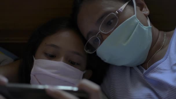 母と娘は保護マスクをしてベッドの上で横になり 娘は携帯電話を使っている コロナウイルス対策コヴィッド19感染症 健康と安全のライフコンセプト — ストック動画