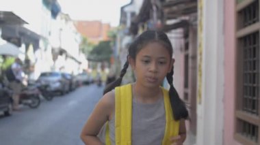 Sırt çantalı Asyalı küçük kız yaz aylarında Phuket 'teki koloni tarzı binaların arasında yaya yolunda rahatça yürüyor. Tayland. Yavaş çekim.