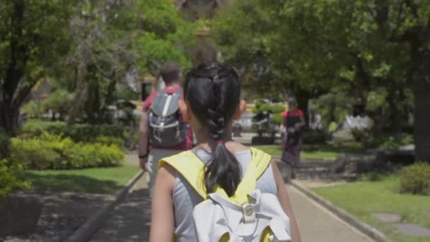 夏の間に多くの観光客と仏教寺院の緑の庭の間の歩道を歩いてバックパックを持つアジアの少女の背面図 スローモーション — ストック動画