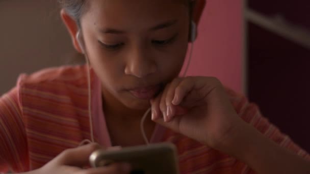 亚洲小女孩一边在家里看网络媒体 一边用带耳机的手机 传播促进娱乐 — 图库视频影像