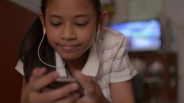 Asyalı tatlı kız yerde yatıyor ve evde sosyal güvenlik için cep telefonunu kullanıyor. Akıllı telefondan müzik videosu izleyen ve kablosuz kulaklıkla dinleyen genç bir kız..