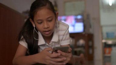 Asyalı tatlı kız yerde yatıyor ve evde sosyal güvenlik için cep telefonunu kullanıyor. Akıllı telefondan müzik videosu izleyen ve kablosuz kulaklıkla dinleyen genç bir kız..