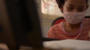 COVID-19 salgını sırasında koruyucu maske takıp evdeki bilgisayarla masaya yazı yazan sevimli bir kız. Ev konseptinde çalış.