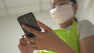 Ev hanımı koruyucu maske takıyor. İletişim kurmak için cep telefonu kullanıyor ve sosyal medyayı evde izliyor. Covid-19 durumu sırasında veri takibi ve cep telefonuyla günlük haberler.