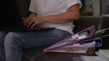 Deri koltukta oturan sıradan bir kadın COVID-19 salgını sırasında evde dizüstü bilgisayarla ve bir yığın belgeyle çalışıyor. Ev kavramından çalış.