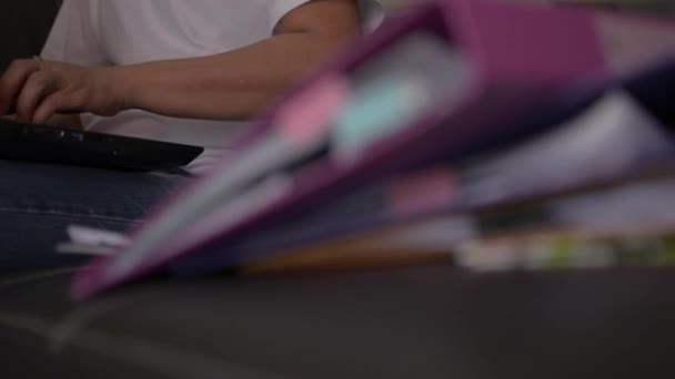 Covid 19発生状況下でノートパソコンを操作する革ソファに腰を下ろして自宅でドキュメントの山を背負ったカジュアルドレス姿の女 家の概念からの仕事 パンニングショット — ストック動画