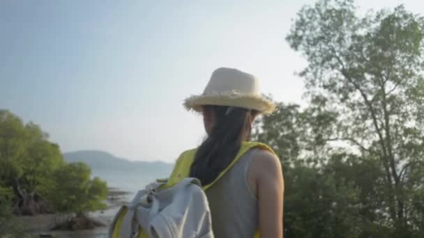 美丽的亚洲姑娘 头戴草帽 背着背包 在夕阳西下的时候 欣赏着美丽的海滨风景 绿树成荫 — 图库视频影像