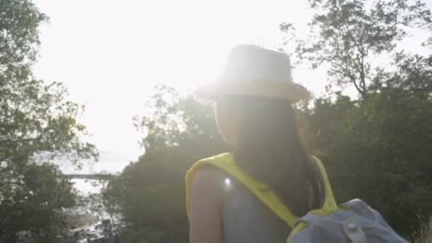 かわいいアジアの女の子は バックパックを歩いてわらの帽子をかぶって 夕暮れ時にマングローブの木で海岸の美しい景色を楽しむ空気に手を上げます スローモーション — ストック動画