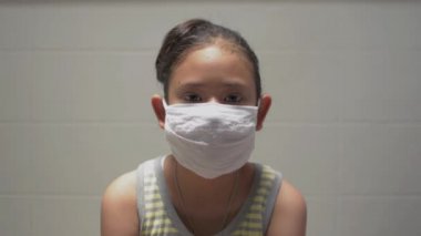 Evde otururken koruyucu maske takan Asyalı şirin bir kız portresi. Genç bir kız, gücünü ve kameraya bakarak gösterir. Sağlık ve motivasyon konsepti. Coronavirus koruması.