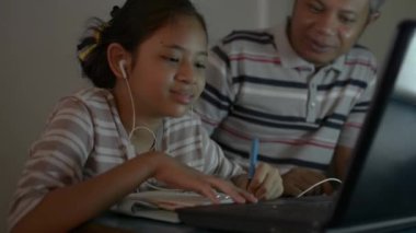 Babası ile birlikte Asyalı tatlı kız ödev yaparken internetten bilgisayar kullanıyor. Babası COVID-19 salgını sırasında kızına evden video aramasıyla yardım edip ders veriyor..