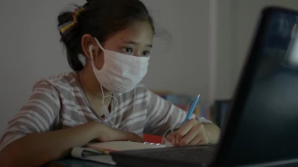 亚洲可爱的女孩在做作业时戴着耳机 在网上看笔记本电脑授课 在Covid 19大流行期间 戴着防护面具的少女在家里带着视频电话学习 — 图库视频影像