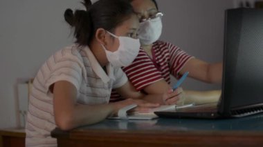 Asyalı tatlı bir kız ve annesi ödev yaparken internette koruyucu maske takıp laptoptan izleme dersi alıyorlar. Anne, COVID-19 salgını sırasında kızına evden video aramasıyla yardım ediyor ve ders veriyor..
