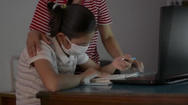 Covid 19パンデミック時に自宅からビデオ通話で勉強する保護マスクを着用したアジア系可愛い女の子 母は立って助けと彼女の娘を教える家庭からのレッスンをオンラインでノートパソコンから — ストック動画