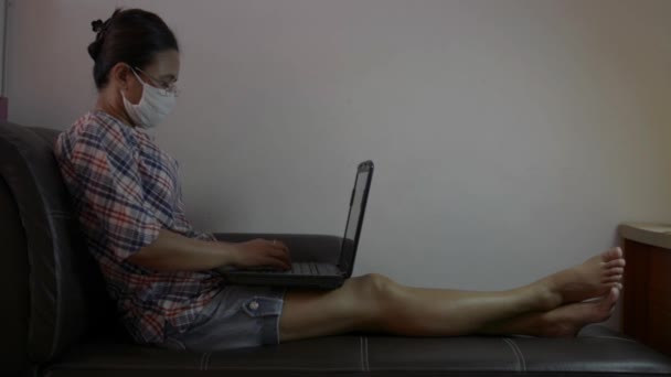 検疫中に自宅から働く保護マスクを身に着けているアジアの女性 Covid 19大流行時のリビングルームでソファに座ってノートパソコンで働くカジュアルな服装のビジネスマン女性 家からの仕事 — ストック動画