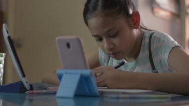 Ev ödevi yaparken cep telefonundan ve dijital tabletten ders izleyen sevimli Asyalı kız. Kadın öğrenci COVID-19 salgını sırasında video ile evden ders çalışıyor..