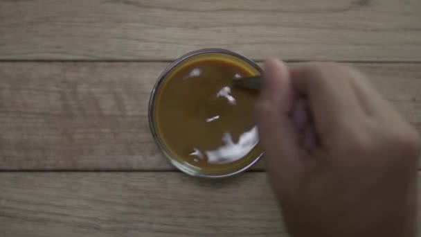 スプーンでカップに熱いコーヒーを攪拌人の手のトップビュー 木製の板の上に練乳と黒コーヒーのカップ背景 — ストック動画