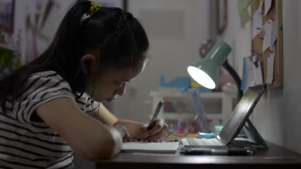 小学生の女の子はイヤフォンを着用し 自宅でオンラインレッスンのためにデジタルタブレットを使用しています Covid 19パンデミックの状況の間にビデオ通話で家から勉強しながら本に書いている女性ティーネージャー — ストック動画
