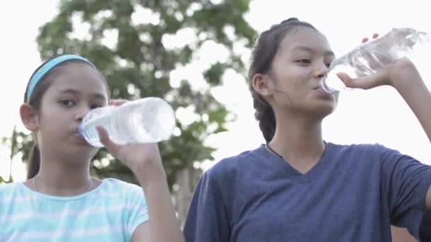 住宅街の公園でペットボトルから水を飲むカジュアルな服装で2人のアジアの可愛い女の子の肖像画 健康な女性のティーンエイジャーと彼女の妹は運動後にミネラルウォーターを飲む スローモーション — ストック動画