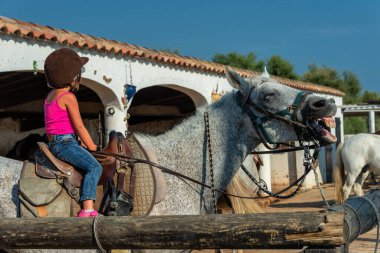  Camargue 'deki bir binicilik merkezinde büyük bir atın üzerinde oturan küçük kız bakıyor ve gülümsüyor.