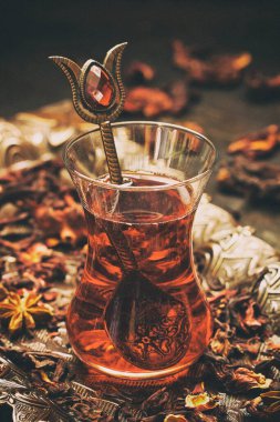 Hibiscus (Karkade) ile bir bardak kırmızı çay, masaya saçılmış yapraklar, koyu arka plan, sığ alan derinliği ile seçici odak