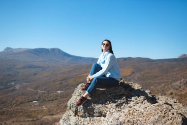 Bir kız dağlarda bir kayada oturuyor ve doğal manzaranın tadını çıkarıyor.