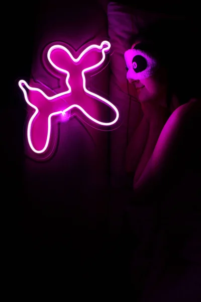 A beautiful girl is sleeping in a sleep blindfold with a purple neon sign dog. Good night. Trendy stylish. Glowing life. Modern style. Neon sign.