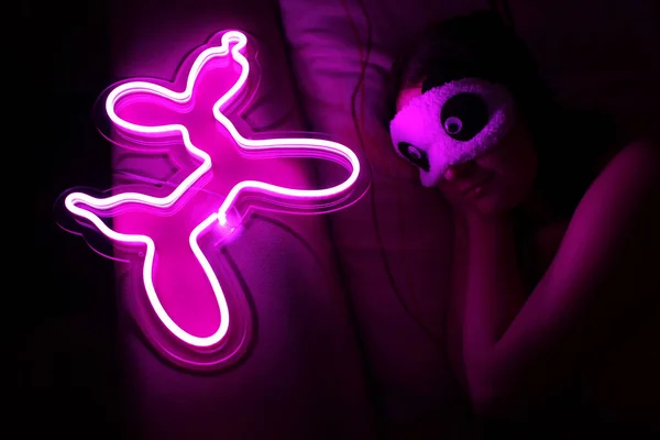 A beautiful girl is sleeping in a sleep blindfold with a purple neon sign dog. Good night. Trendy stylish. Glowing life. Modern style. Neon sign.