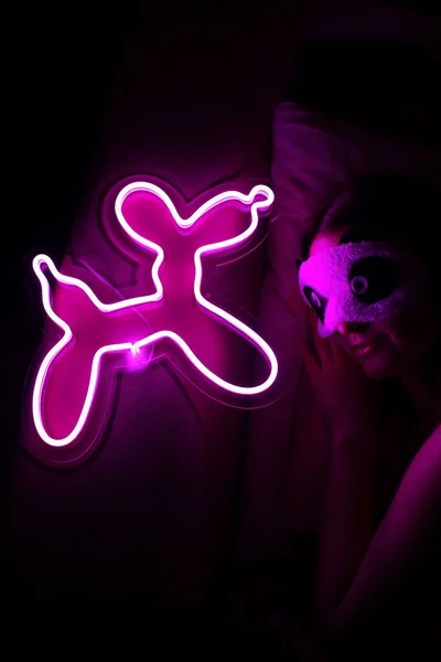 A beautiful girl is sleeping in a sleep blindfold with a purple neon sign dog. Good night. Trendy stylish. Glowing life. Modern style. Neon sign.