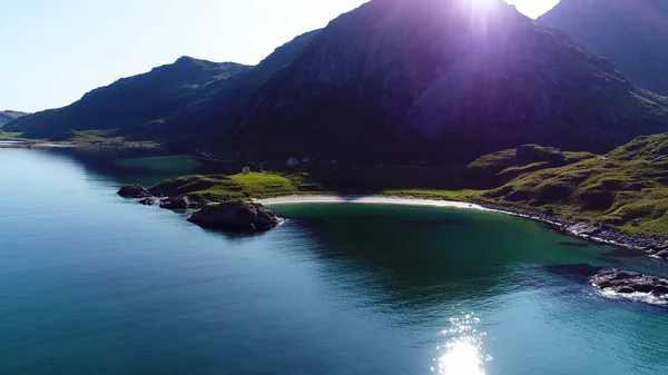 The sun breaks through the top of a mountain in Norway, at the foot of which is the beach near the fjord