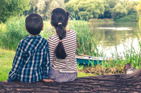 Két gyerek, fiú és lány, ülnek a fatörzsön és nézik a tavi tájat. Stock Kép