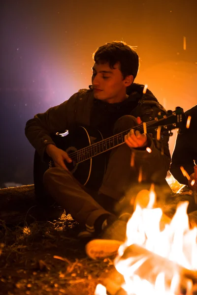 Tipo tocando la guitarra alrededor de una fogata en el fondo del cielo estrellado — Foto de Stock