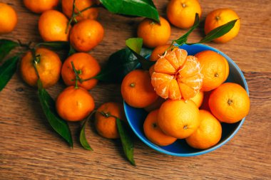 Ahşap bir masanın üzerinde duran mavi kasede bir sürü taze mandalina var. 