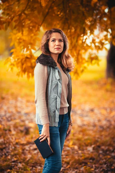 Retrato de uma jovem em colete de peles e livro de leitura no fundo do parque de outono — Fotografia de Stock