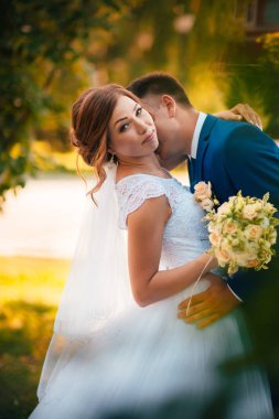 Gelin ve damat, doğa parkında sarılıp öpüşüyor.