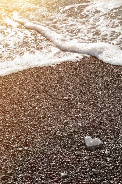一个灰色的石头心形的海岸与海洋泡沫和清澈的水在日落的黑色沙滩上。 爱的概念，婚礼，自由。 带有复制空间的情人节贺卡的背景. — 图库照片