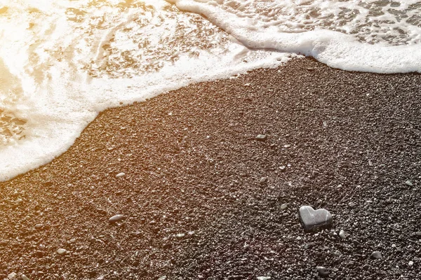 海岸上躺着一块灰色的、心形的石子，在日落的时候，在黑色的沙滩上漂浮着泡沫和清澈的水。 爱的概念，婚礼，自由。 带有复制空间的情人节贺卡的背景. — 图库照片