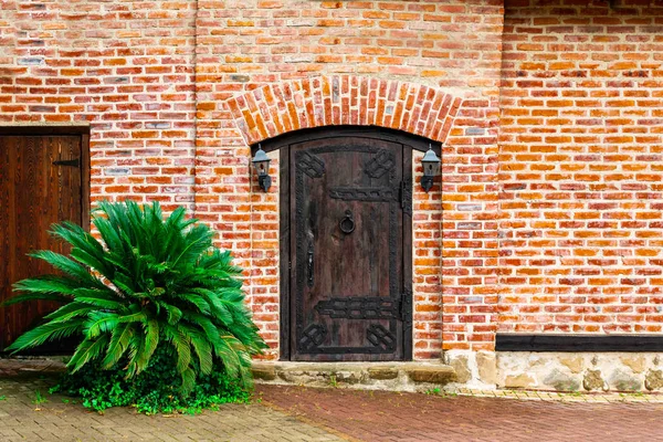 Une vieille porte massive en bois fermée avec une poignée de porte en forme d'anneau torsadé, des lanternes vintage suspendues des deux côtés d'un mur de briques rouges et un arbuste vert. Contexte, espace de copie . — Photo